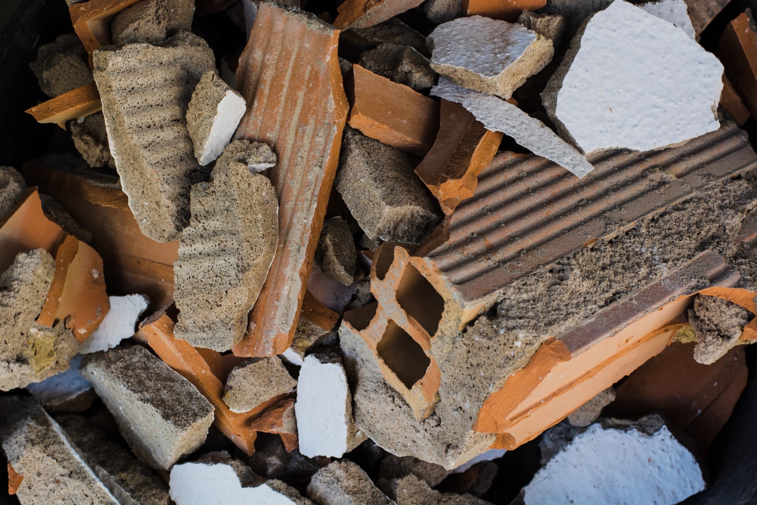 A close-up of mixed construction debris including broken bricks, tiles, and plaster fragments in a dark container.